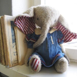 May include: A plush elephant toy wearing a blue dress with white buttons and a red and white striped scarf. The toy has a long trunk and is sitting on a white surface.