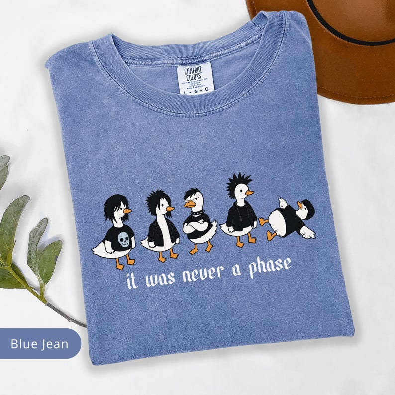 Puede incluir: Camiseta azul vaquero con cuello redondo. La camiseta presenta un gr&aacute;fico de cinco patos de dibujos animados vestidos con atuendos alternativos, con el texto "it was never a phase" debajo.