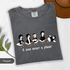 May include: Pepper-colored Comfort Colors t-shirt featuring a graphic of five cartoon ducks in punk attire. The ducks are in various poses, with the text "it was never a phase" below them. The shirt has a crew neck and short sleeves.