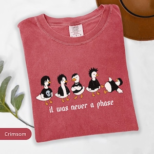 Puede incluir: Camiseta de color carmes&iacute; con un gr&aacute;fico de cinco patos de dibujos animados con atuendos alternativos. Los patos tienen diferentes peinados y ropa, con el texto "it was never a phase" debajo. La camiseta tiene cuello redondo.