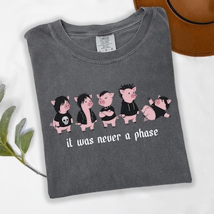 May include: A charcoal gray t-shirt featuring a graphic of five cartoon pigs dressed in punk-inspired clothing. Below the pigs, the text reads "it was never a phase" in a white gothic-style font. The shirt has a crew neck and a Comfort Colors label.