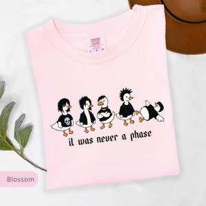 Puede incluir: Camiseta rosa claro con un gr&aacute;fico de cinco patos de dibujos animados vestidos con atuendos punk rock. El texto "it was never a phase" est&aacute; impreso debajo. La camiseta tiene cuello redondo y la etiqueta de la marca "Comfort Colors" es visible.