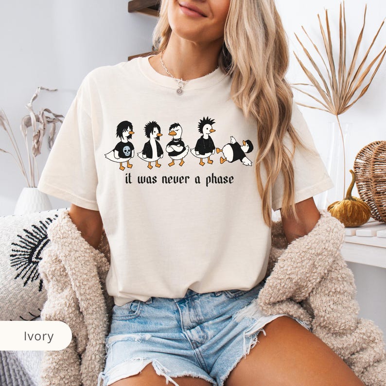 Puede incluir: Camiseta de color marfil con un gr&aacute;fico de cinco patos de dibujos animados con peinados y ropa punk en blanco y negro. El texto "it was never a phase" est&aacute; impreso debajo en una fuente g&oacute;tica. La camiseta tiene un corte holgado.