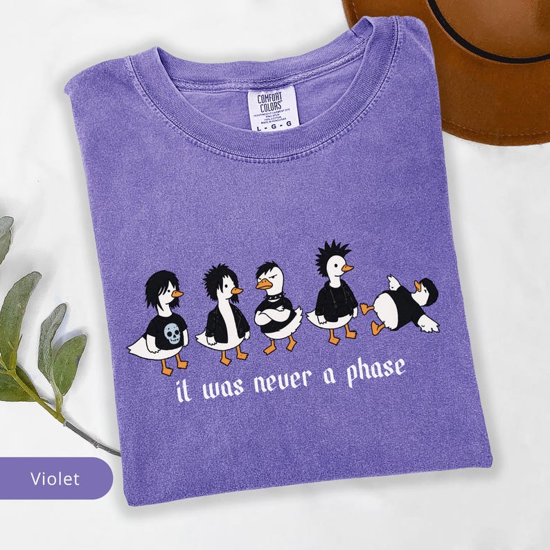 Puede incluir: Camiseta violeta Comfort Colors con un gr&aacute;fico de cinco patos de dibujos animados con ropa alternativa. Los patos llevan chaquetas negras y camisetas con un gr&aacute;fico de calavera. El texto "it was never a phase" est&aacute; impreso debajo.