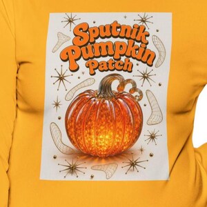 May include: A long-sleeve shirt in mustard yellow featuring a graphic print on a white rectangular background. The print displays the text "Sputnik Pumpkin Patch" in orange, a central orange pumpkin, and surrounding wireframe shapes and starbursts.