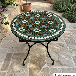 Puede incluir: Mesa redonda de mosaico con estructura de metal negro. La superficie presenta un patrón geométrico en verde, marrón, blanco y azul. La mesa está al aire libre, rodeada de plantas y suelo de piedra. Diámetro aproximado de 75 cm.