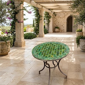 Puede incluir: Mesa redonda de jardín con tablero de mosaico verde. La mesa tiene una base de hierro forjado. La mesa tiene un diámetro de aproximadamente 60 cm. En el fondo, una pérgola y un edificio de piedra.