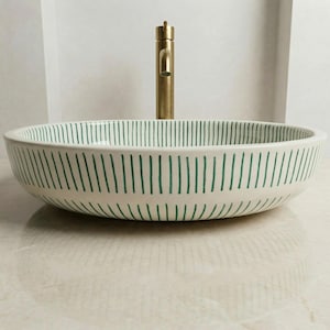 May include: A white ceramic vessel sink with a green vertical stripe pattern. A gold-colored faucet is mounted behind the sink. The sink is round and sits on a light-colored surface.