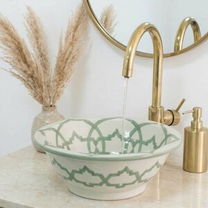 Puede incluir: Un lavabo de cerámica blanca con un patrón geométrico verde. Se combina con un grifo y un dispensador de jabón dorados. Un jarrón de hierba de la pampa seca y un espejo dorado están en el fondo.