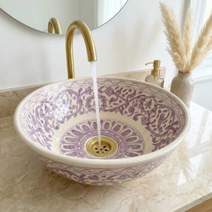 May include: A decorative, round ceramic sink with a floral pattern in purple and white. A gold faucet pours water into the sink. The sink is on a marble countertop, with a mirror and decorative items in the background.