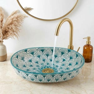 Op de afbeelding: Een decoratieve keramische wastafel met een turquoise en wit bloemmotief. De ronde wastafel heeft een gouden afvoer en is gemonteerd op een marmeren aanrecht. Een gouden kraan bevindt zich erboven, waar water uit stroomt. Rechts staat een bruine zeepdispenser.