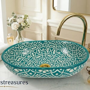 Puede incluir: Lavabo ovalado de cerámica con un patrón geométrico turquesa y blanco. El lavabo está sobre una encimera blanca con un grifo dorado. La imagen también muestra un ramo de flores blancas, toallas y objetos decorativos.