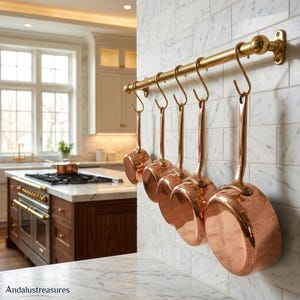 May include: Copper saucepans with long handles hanging from a brass rail in a kitchen. The pans are displayed against a white tiled wall. The kitchen has a marble countertop and a stainless steel stove.
