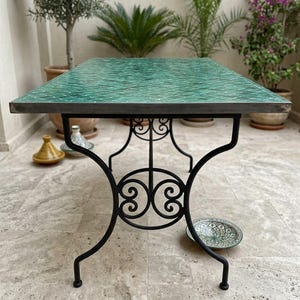 May include: A rectangular table featuring a green mosaic tile top with a diamond pattern and a black wrought iron base with decorative scrollwork. The table has a metal frame edge and stands on four curved legs with a central circular support.