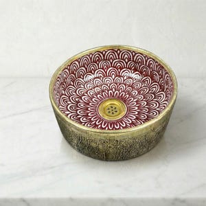 May include: A decorative ceramic sink with a burgundy and white floral pattern inside. The sink has a gold-colored rim and a textured, aged bronze exterior. The drain is gold-colored.