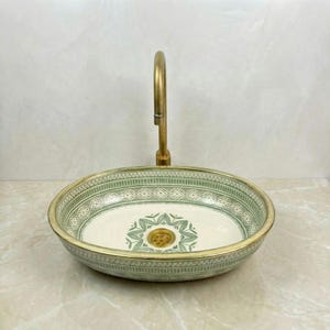 May include: A round ceramic sink with a brass tap. The sink has a white interior with a green floral design and a green patterned border. The tap is a curved, single-handle design, adding a touch of elegance.