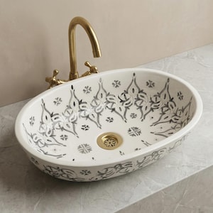 Puede incluir: Lavabo ovalado de cerámica blanca con un patrón floral decorativo en negro y gris. El lavabo tiene un desagüe dorado y se combina con un grifo dorado.