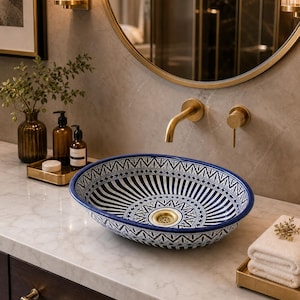 May include: An oval ceramic vessel sink featuring a blue and white geometric pattern with a central gold-toned drain. The sink is positioned on a white marble countertop next to a stack of white towels and amber glass bottles.