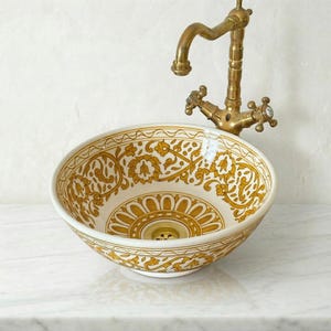 Puede incluir: Un lavabo de cerámica decorativo con un diseño floral dorado sobre fondo blanco. El lavabo se combina con un grifo de latón de estilo vintage. El lavabo es redondo y tiene un desagüe central. El diámetro del lavabo es de aproximadamente 30 cm.