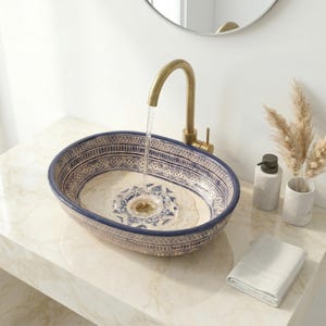 May include: An oval ceramic bathroom sink with a blue and white patterned design. A gold faucet pours water into the sink. A white marble countertop holds the sink, a soap dispenser, a vase with dried plants, and a folded white towel.