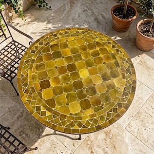 May include: A round table featuring a mosaic top composed of numerous octagonal and diamond-shaped tiles in varying shades of olive green and mustard yellow. The table has a dark metal frame and the text Andalustreasures is visible at the bottom.