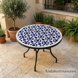 Puede incluir: Mesa redonda de exterior con una tapa de mosaico con un patrón de estrellas azules y blancas. La mesa tiene una estructura y patas de metal oscuro. La mesa tiene aproximadamente 61 cm de diámetro.