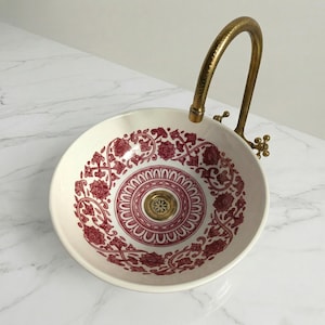 Könnte beinhalten: Ein dekoratives, rundes Keramikwaschbecken mit einem rot-weißen Blumenmuster. Das Waschbecken hat einen Wasserhahn und einen Abfluss aus Messing. Das florale Design ist um den Abfluss zentriert und von einem sich wiederholenden Muster umgeben.