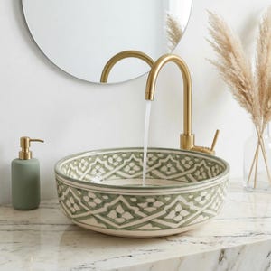 Puede incluir: Un lavabo de cerámica decorativo con un diseño floral verde y blanco. Un grifo dorado vierte agua en el lavabo. Un dispensador de jabón verde y un jarrón con plantas secas están en la encimera.