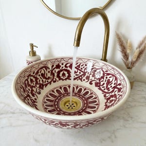Puede incluir: Un lavabo de cerámica redondo con base color crema y patrones florales y geométricos en rojo oscuro. Un grifo de latón alto y curvo se extiende sobre el cuenco, con un flujo constante de agua cayendo hacia el desagüe dorado.