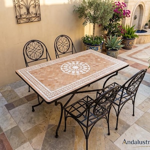 Può includere: Un tavolo da pranzo rettangolare per esterni con un piano in mosaico color beige e bianco caratterizzato da un motivo geometrico centrale e una struttura in metallo nero, accompagnato da quattro sedie in metallo nero con schienali a volute.
