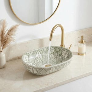 Pode incluir: Lavatório oval de cerâmica com um padrão decorativo verde e branco. Uma torneira dourada está a deitar água para dentro do lavatório. Um espelho com moldura dourada e um dispensador de sabonete são visíveis no fundo.