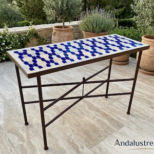 Puede incluir: Una mesa consola rectangular con un tablero de azulejos de mosaico que presenta un patrón geométrico repetitivo en colores azul oscuro y crema. La mesa está sostenida por una estructura de metal marrón oscuro con cuatro patas rectas y un refuerzo en cruz.
