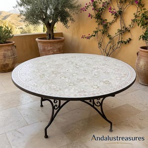 Puede incluir: Mesa redonda de mosaico para exteriores con un patrón decorativo en tonos blancos y beige. La mesa tiene una estructura y patas de metal oscuro con detalles en espiral. La mesa tiene aproximadamente 122 cm de diámetro.