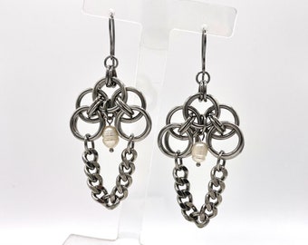 Pode incluir: Um par de brincos de prata com uma corrente e um pingente de pérola. Os brincos estão em um suporte transparente com uma concha branca e pérolas abaixo.
