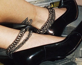 Pode incluir: Sapatos pretos de salto alto com bico fino e detalhe de corrente no tornozelo. Os sapatos têm um acabamento brilhante. Uma tatuagem é visível no tornozelo. Os sapatos estão sobre uma superfície metálica.