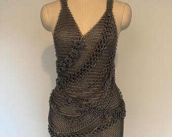Pode incluir: Um vestido de cota de malha prateado com decote em V e alças finas. O vestido é exibido em um manequim e apresenta detalhes intrincados e drapeado de corrente. O vestido de metal é uma peça de roupa única.