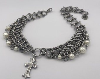 Pode incluir: Um bracelete de corrente prateada com um fecho de cruz e detalhes de pérolas falsas.