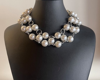 Pode incluir: Um colar de pérolas em camadas com pérolas grandes, redondas e brancas. As pérolas são unidas por elos de metal prateado. O colar é exibido em um busto de manequim preto sobre um fundo neutro.