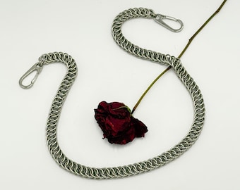 Pode incluir: Um colar de malha de corrente prateado com dois fechos de mosquetão, sobre um fundo branco. Uma rosa vermelha escura seca e seu caule são colocados perto do colar.