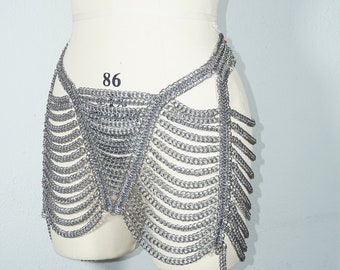 Pode incluir: Uma saia de malha de corrente prateada com um design único. A saia é feita de anéis de metal interligados e tem um visual fluido e em camadas. A saia é exibida em um manequim branco.