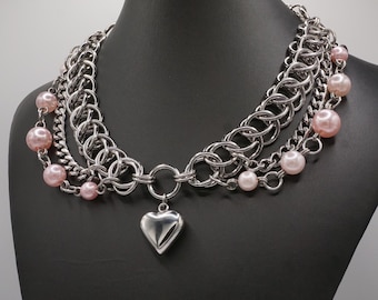 Pode incluir: Um colar de corrente prateada em camadas com um pingente de coração e detalhes de pérolas cor-de-rosa.