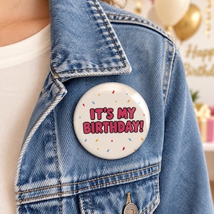 May include: A circular pinback button featuring a white background with colorful sprinkles and the text IT'S MY BIRTHDAY! in bold pink letters. The button is attached to a blue denim jacket with visible stitching and a metal button detail.