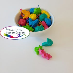 Puede incluir: Un cuenco blanco lleno de ceras de colores con formas únicas. Las ceras tienen varias formas, incluyendo mariposas, caracoles y peces, en tonos amarillos, rosas, azules y verdes. El logotipo de The Kids Table Custom Crayons es visible.