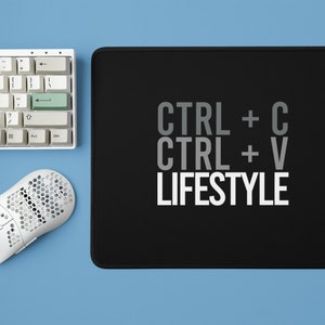 May include: A black mouse pad with white text that reads "CTRL + C CTRL + V LIFESTYLE". A white keyboard and a white mouse are on a blue surface.