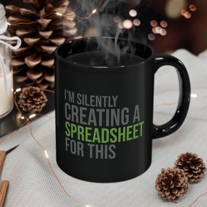 May include: Black coffee mug with white text that reads "I'm silently creating a spreadsheet for this". The text "creating a spreadsheet" is in green. The mug is sitting on a light brown surface with a string of lights.