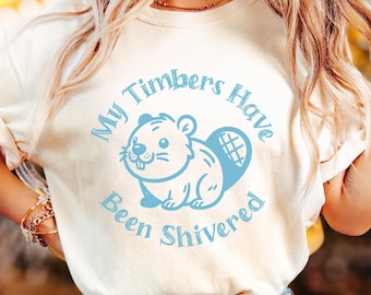 Peut inclure: Un t-shirt blanc avec un graphique bleu d'un castor et le texte "My Timbers Have Been Shivered". Le t-shirt est étiqueté "IVORY" dans le coin inférieur gauche.