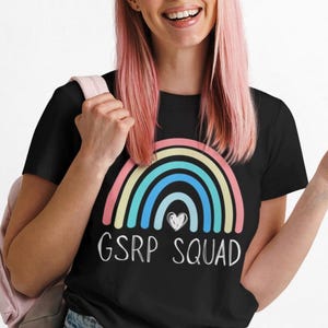 May include: Black crew neck t-shirt with a rainbow design and the text "GSRP SQUAD". The rainbow features pastel colors. The person in the image is wearing blue jeans and a pink backpack.
