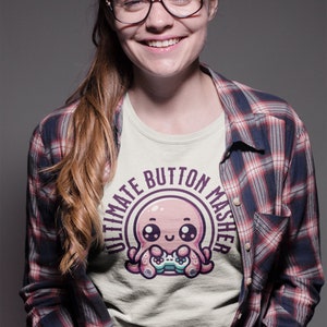 May include: A woman wearing a light beige t-shirt with a pink octopus holding a video game controller. The octopus is surrounded by a purple circle with the text "Ultimate Button Masher". She is wearing a red and black plaid shirt over the t-shirt.