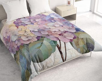 Può includere: Un copriletto con una stampa floreale su larga scala di fiori di ortensia in tonalità rosa tenue, lavanda e blu, accentuata da foglie verdi e steli marrone scuro, abbinato a due cuscini quadrati bianchi a tinta unita sul letto.