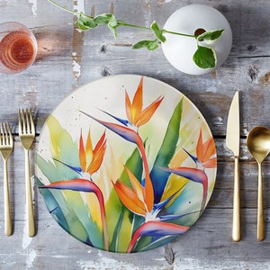Pu&ograve; includere: Un piatto rotondo bianco con un disegno ad acquerello di fiori di uccello del paradiso arancioni, gialli, blu e verdi. Il piatto &egrave; su un tavolo di legno con posate d'oro.
